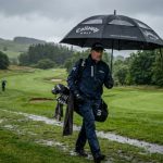 A golfer holding a large heavy-duty golf umbrella with a vented canopy on a rainy golf course fairway