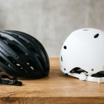 A matte black bicycle helmet and white skateboard helmet sitting side by side on a wooden table.