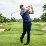 Golfer wearing a navy golf vest completes a swing on a green course.